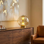 Cognac-tinted glass sphere of this Clérachic lamp lit on a walnut sideboard, with a gold-flecked abstract painting behind.