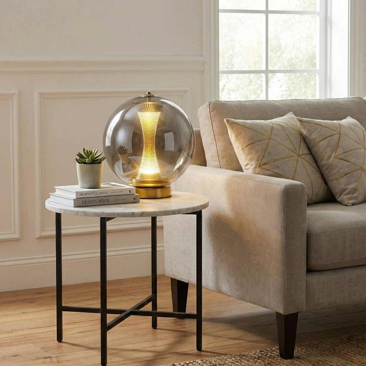Clear glass sphere of this Clérachic lamp on a marble-topped side table by a cream sofa, with a small plant and a stack of books.