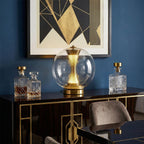 This Clérachic sphere lamp lit on a black and gold cabinet, against a navy wall with a geometric gold artwork and crystal decanters.