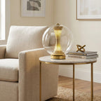 Clear-glass Clérachic sphere lamp lit on a marble and brass side table beside a cream armchair, with books and a gold ornament.