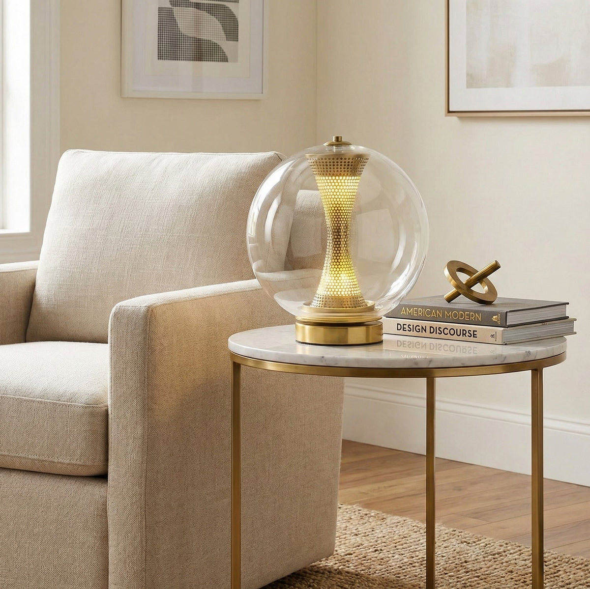 Clear-glass Clérachic sphere lamp lit on a marble and brass side table beside a cream armchair, with books and a gold ornament.