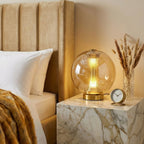 A Clérachic cognac sphere lamp lit on a marble-look bedside block, with a headboard, dried pampas stems and a small clock.