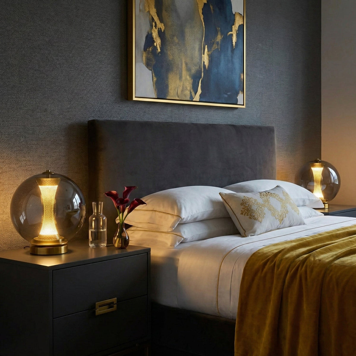 Two Clérachic smoky grey sphere lamps lit either side of a made bed with a mustard throw and a dark grey headboard.