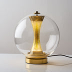 Front view of this Clérachic clear-glass sphere lamp, lit, its perforated brass column glowing above a gold base with a black cord.