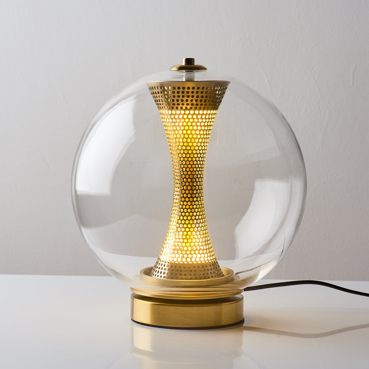 Front view of this Clérachic clear-glass sphere lamp, lit, its perforated brass column glowing above a gold base with a black cord.
