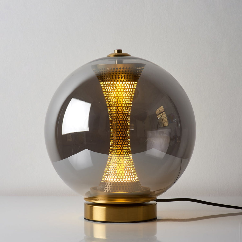 Smoky grey glass sphere of this Clérachic lamp, lit on a white surface, with the perforated brass column glowing inside.