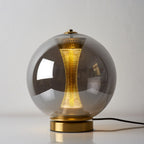 Smoky grey glass sphere of this Clérachic lamp, lit on a white surface, with the perforated brass column glowing inside.