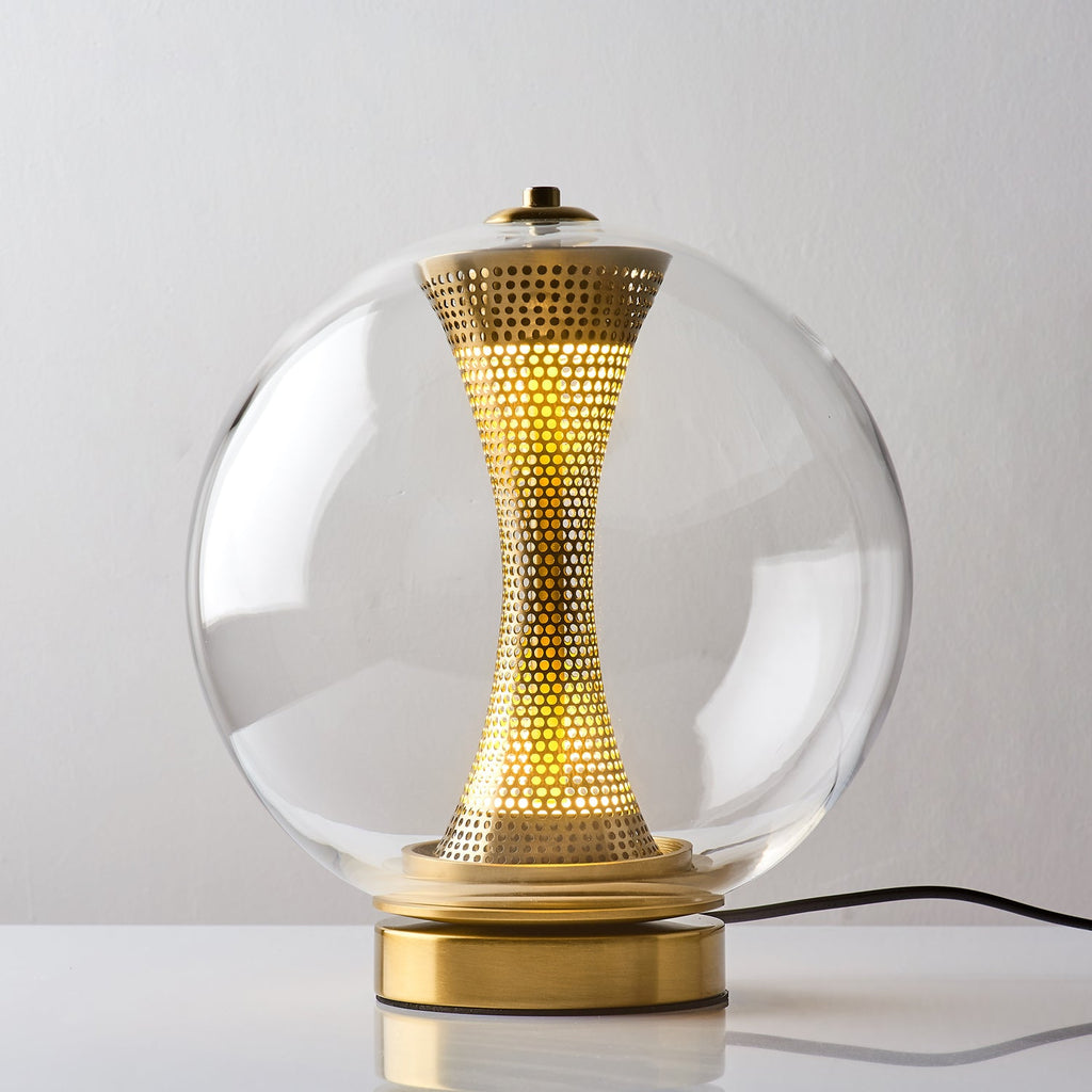 This Clérachic clear-glass sphere lamp, lit, on a white table with the black cord running behind its gold base.