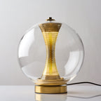 This Clérachic clear-glass sphere lamp, lit, on a white table with the black cord running behind its gold base.