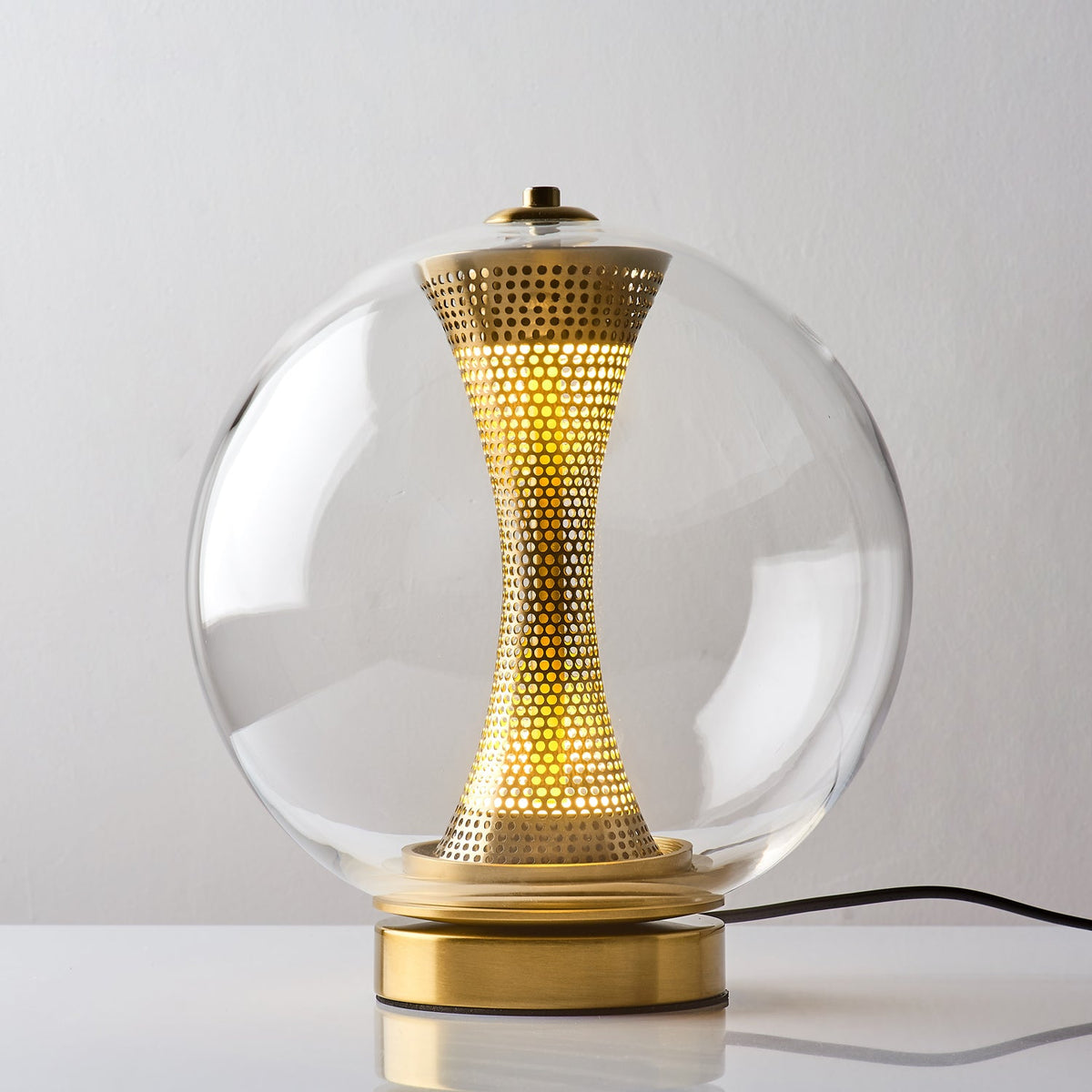 This Clérachic clear-glass sphere lamp, lit, on a white table with the black cord running behind its gold base.