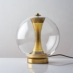 This Clérachic clear-glass sphere lamp lit on a white surface, a warm glow showing through the holes in the brass column.