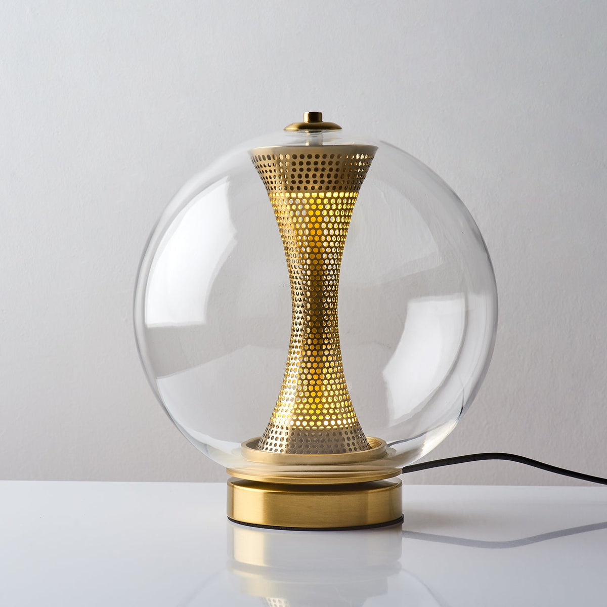 This Clérachic clear-glass sphere lamp lit on a white surface, a warm glow showing through the holes in the brass column.