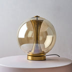Compact Clérachic cognac sphere lamp on a pale round stand, unlit, with the brass column visible through the tinted glass.