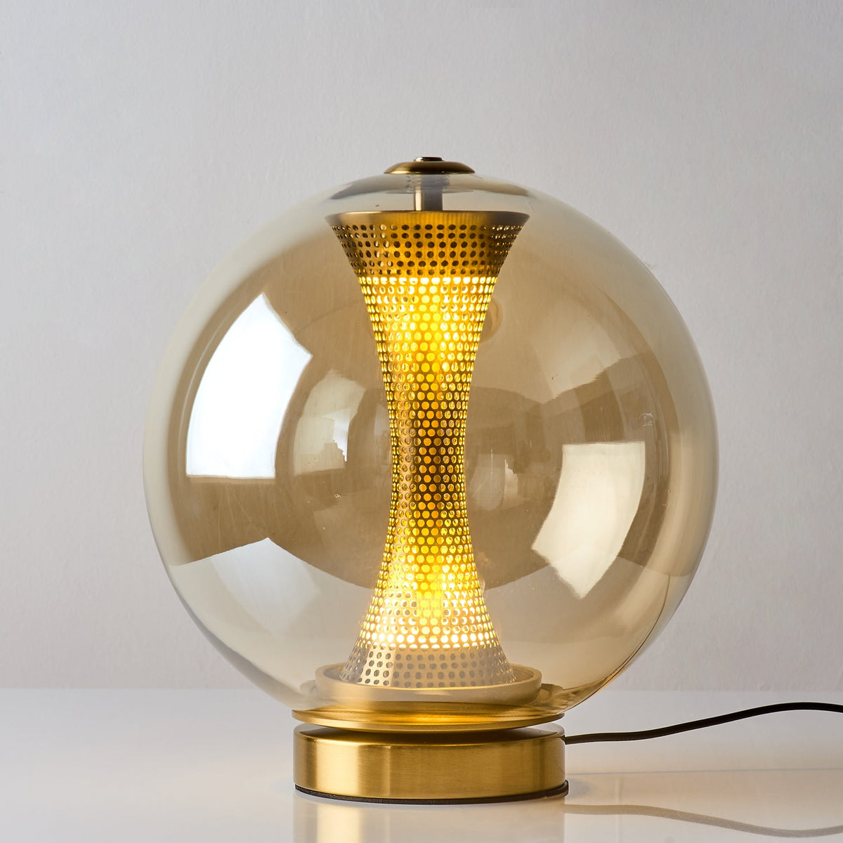 Lit Clérachic cognac glass sphere lamp on white, a yellow glow showing through the perforated brass column and the amber glass.