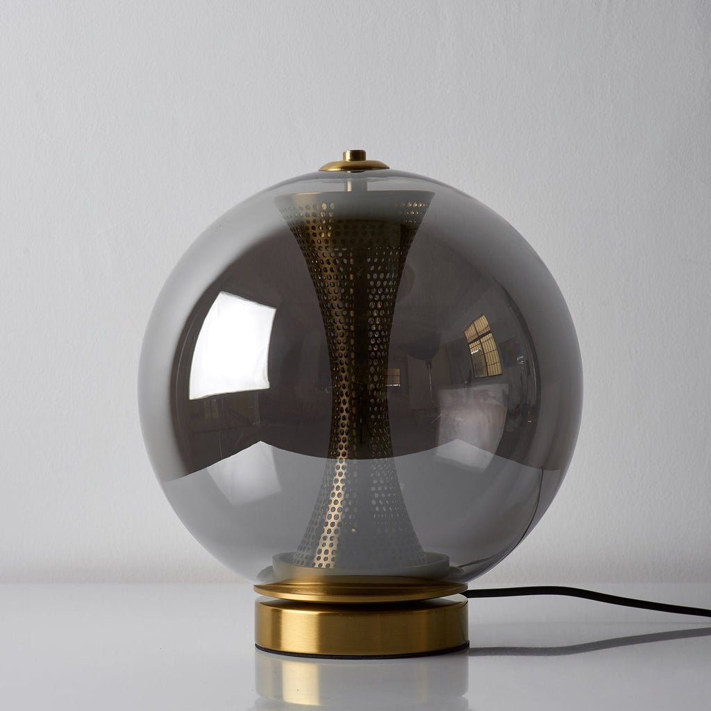 Unlit Clérachic smoky grey sphere lamp on a white surface, the dark glass hiding most of the perforated brass column.