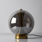 Unlit Clérachic smoky grey sphere lamp on a white surface, the dark glass hiding most of the perforated brass column.