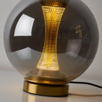 Close view of the lit column through the smoky grey glass of this Clérachic lamp, with the gold base and black cord below.
