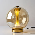 Lit Clérachic cognac sphere lamp on a white surface, its amber glass and glowing brass column above a gold base.