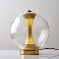 Front view of this lit Clérachic clear-glass sphere lamp, a soft glow in the perforated column above the gold base.