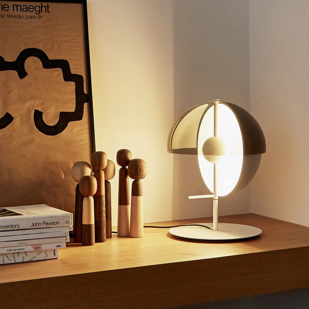 On a wooden shelf beside carved figures and books, this white-based Clérachic table lamp glows warm through its dome and half-sphere.