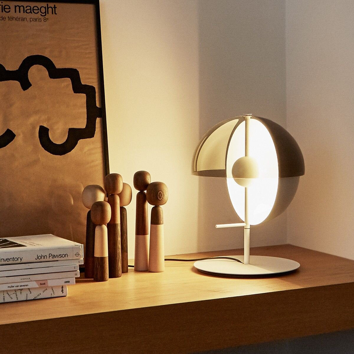 On a wooden shelf beside carved figures and books, this white-based Clérachic table lamp glows warm through its dome and half-sphere.