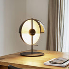A black-based Clérachic table lamp lit on an oak desk beside a leather notebook, with linen curtains hanging behind it.