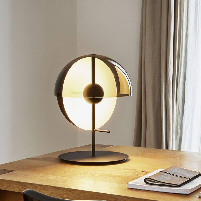 A black-based Clérachic table lamp lit on an oak desk beside a leather notebook, with linen curtains hanging behind it.