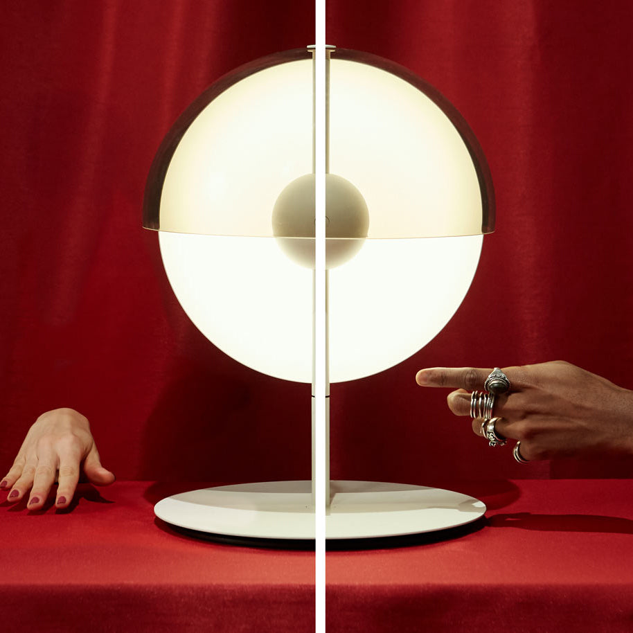 Close view of a white-based Clérachic table lamp against red fabric, with its glowing half-sphere and central ball in focus.