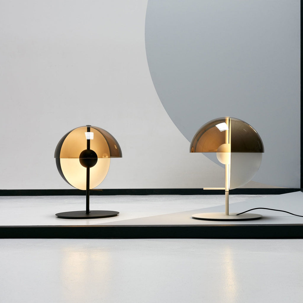 One black and one white Clérachic table lamp stand side by side on a pale studio floor against a grey backdrop.