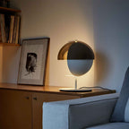 A white-based Clérachic table lamp on a wood sideboard beside a framed ink print, with the shade turned to the wall.