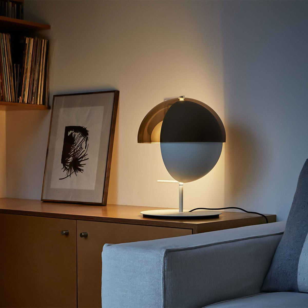 A white-based Clérachic table lamp on a wood sideboard beside a framed ink print, with the shade turned to the wall.