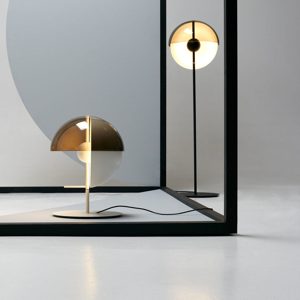 In a pale studio, a white-based Clérachic table lamp stands in front of a black frame, with a tall floor lamp reflected behind.