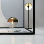 In a pale studio, a white-based Clérachic table lamp stands in front of a black frame, with a tall floor lamp reflected behind.