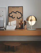 A white-based Clérachic table lamp stands on a wood shelf with carved figures and a framed poster, in soft daylight.