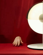 Against a red fabric backdrop, a hand rests on the table beside the glowing half-sphere of a white Clérachic table lamp.