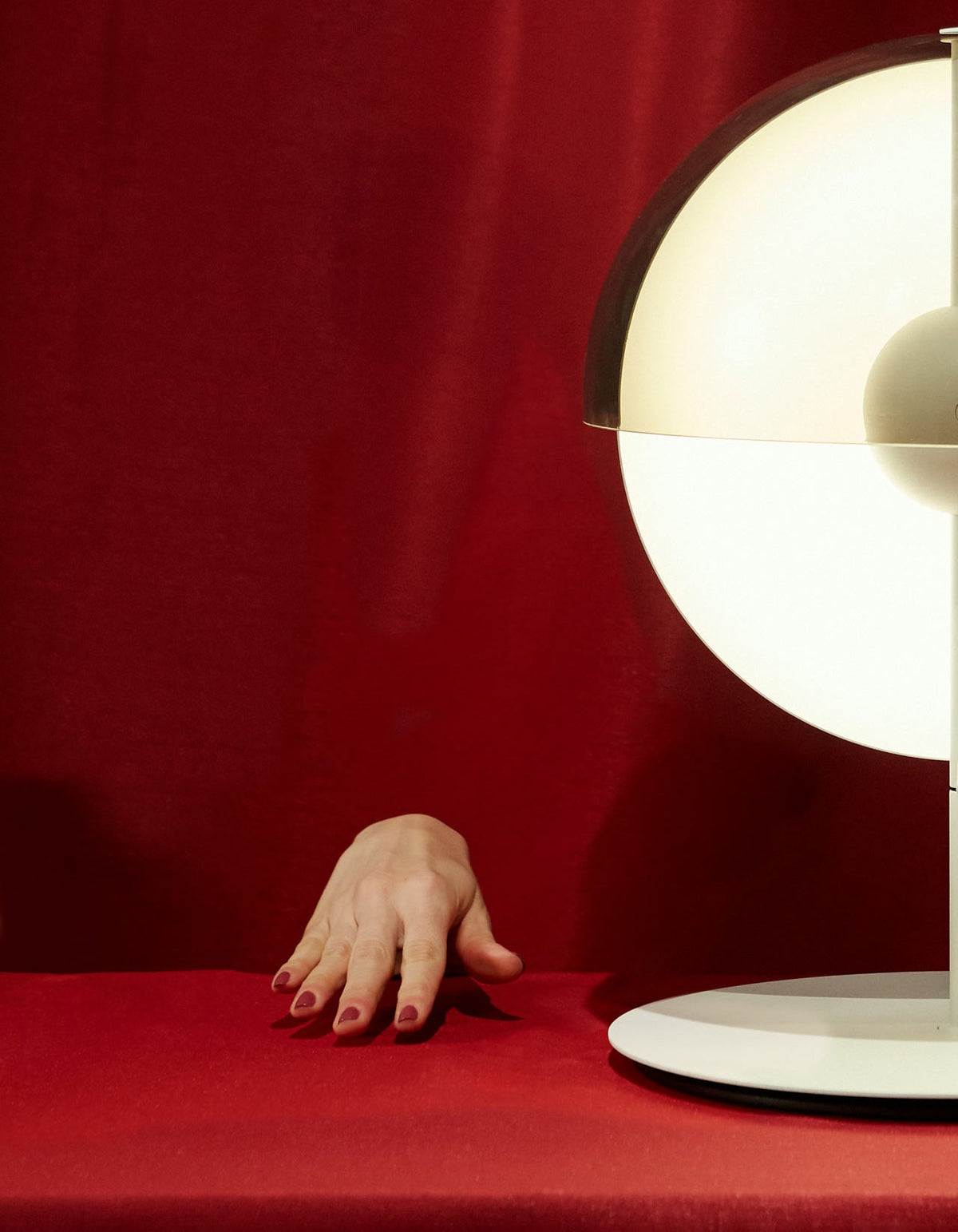 Against a red fabric backdrop, a hand rests on the table beside the glowing half-sphere of a white Clérachic table lamp.