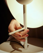 A hand holds the adjustable white stem of this Clérachic table lamp, its glass half-sphere glowing above on a dark ground.