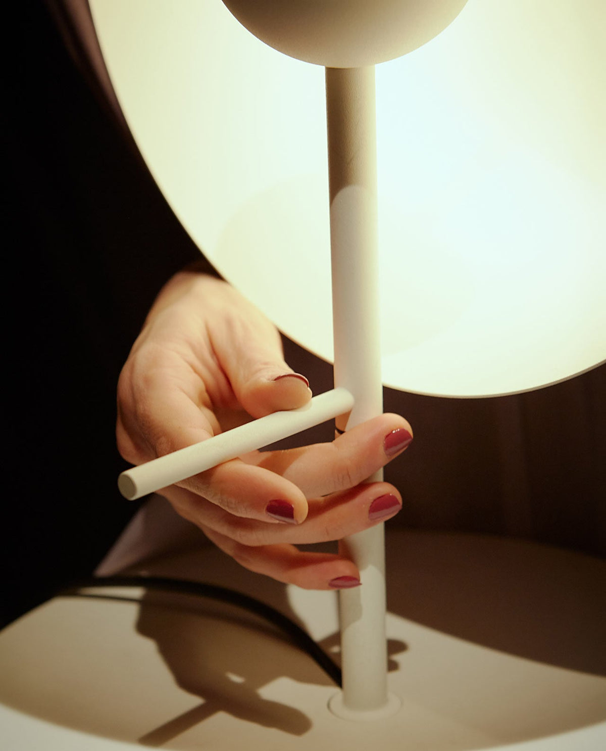 A hand holds the adjustable white stem of this Clérachic table lamp, its glass half-sphere glowing above on a dark ground.