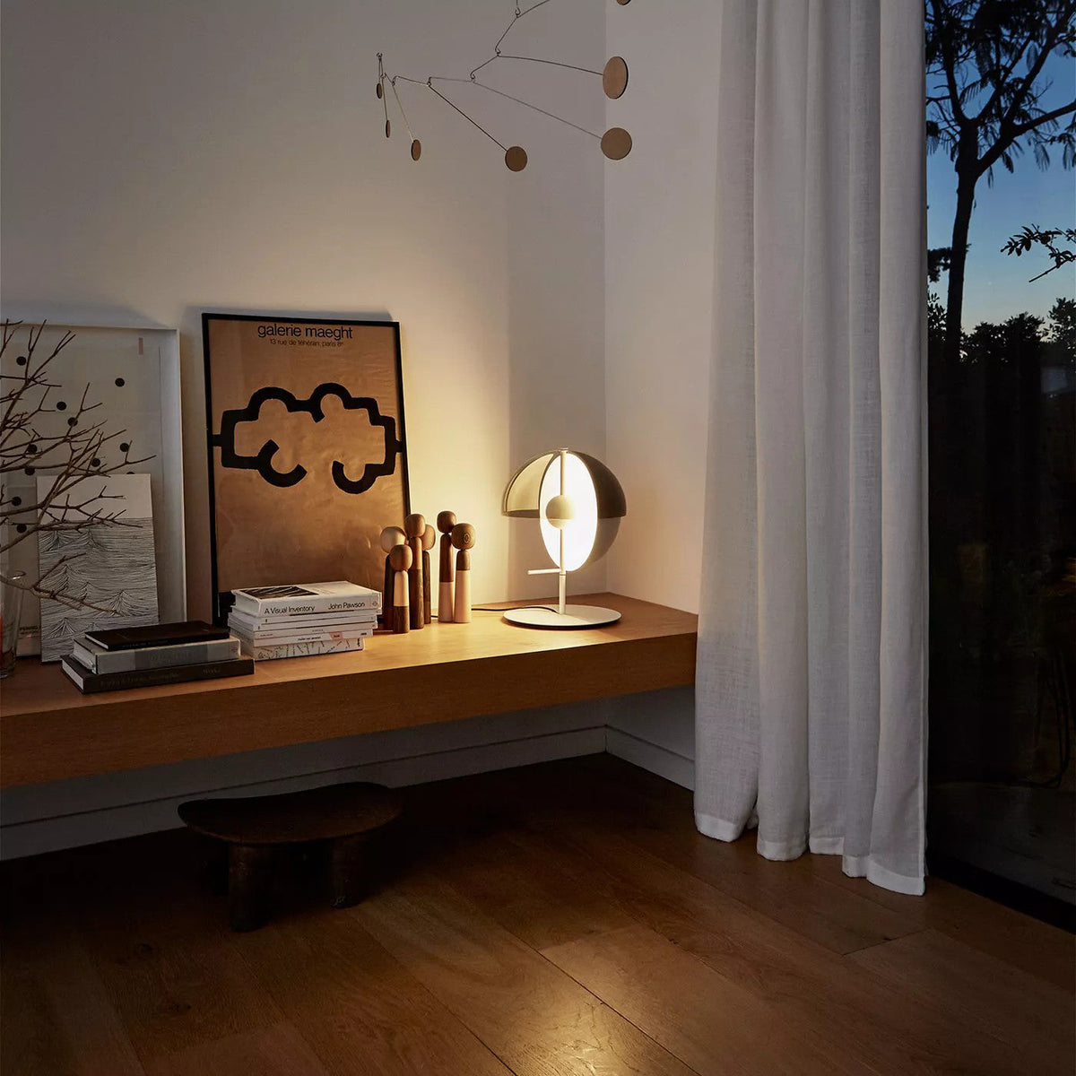 Lit at dusk, a white-based Clérachic table lamp sits on a wooden shelf by a window with white curtains and framed art.