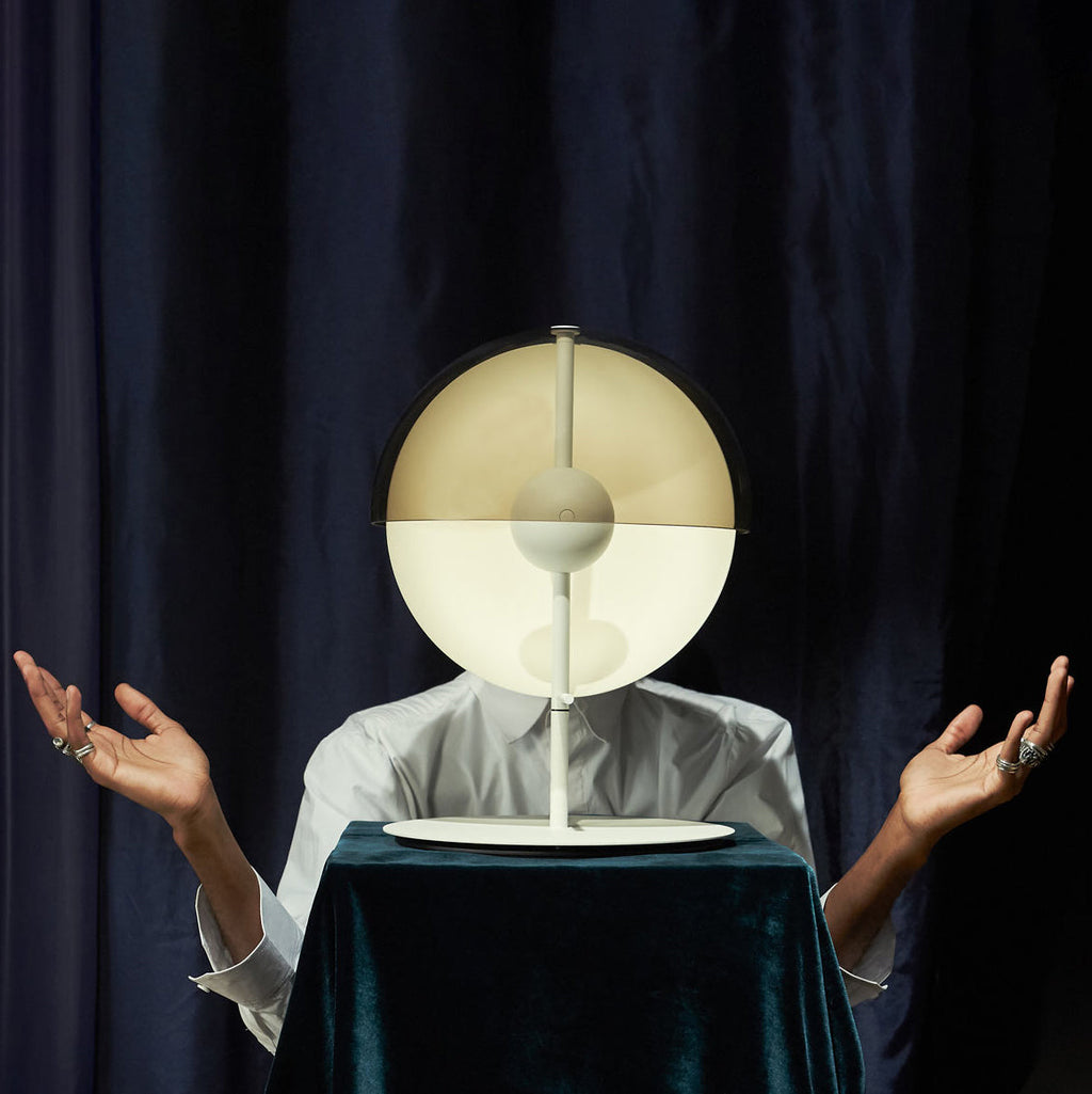 A person's face is hidden behind a white-based Clérachic table lamp held on a draped table before a navy curtain.