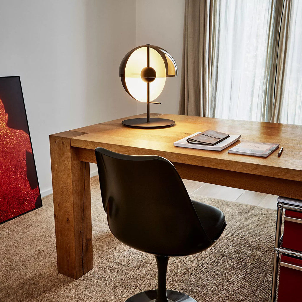 A black-based Clérachic table lamp lit on a solid oak desk beside notebooks, a black swivel chair and a linen curtain.