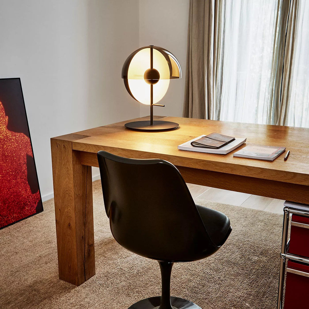A black-based Clérachic table lamp lit on a solid oak desk beside notebooks, a black swivel chair and a linen curtain.