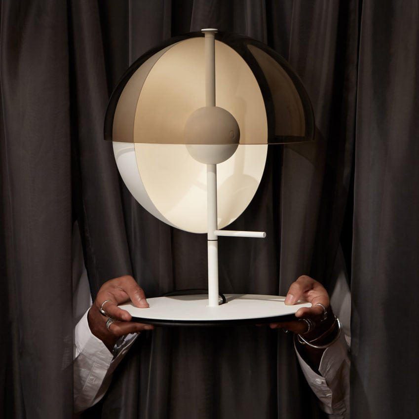 A pair of hands holds a white-based Clérachic table lamp in front of a dark grey curtain, the half-dome tilted forward.