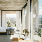 On a timber bedside shelf in a concrete room, a white-based Clérachic table lamp looks out over a bed and a view of hills.