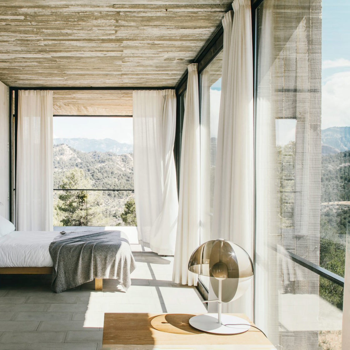 On a timber bedside shelf in a concrete room, a white-based Clérachic table lamp looks out over a bed and a view of hills.
