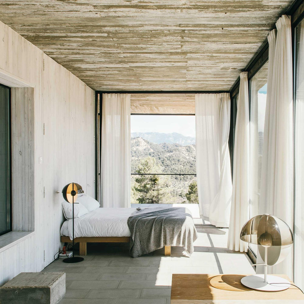 In a concrete-walled bedroom with a view of hills, a white-based Clérachic table lamp stands in the lower right of the frame.