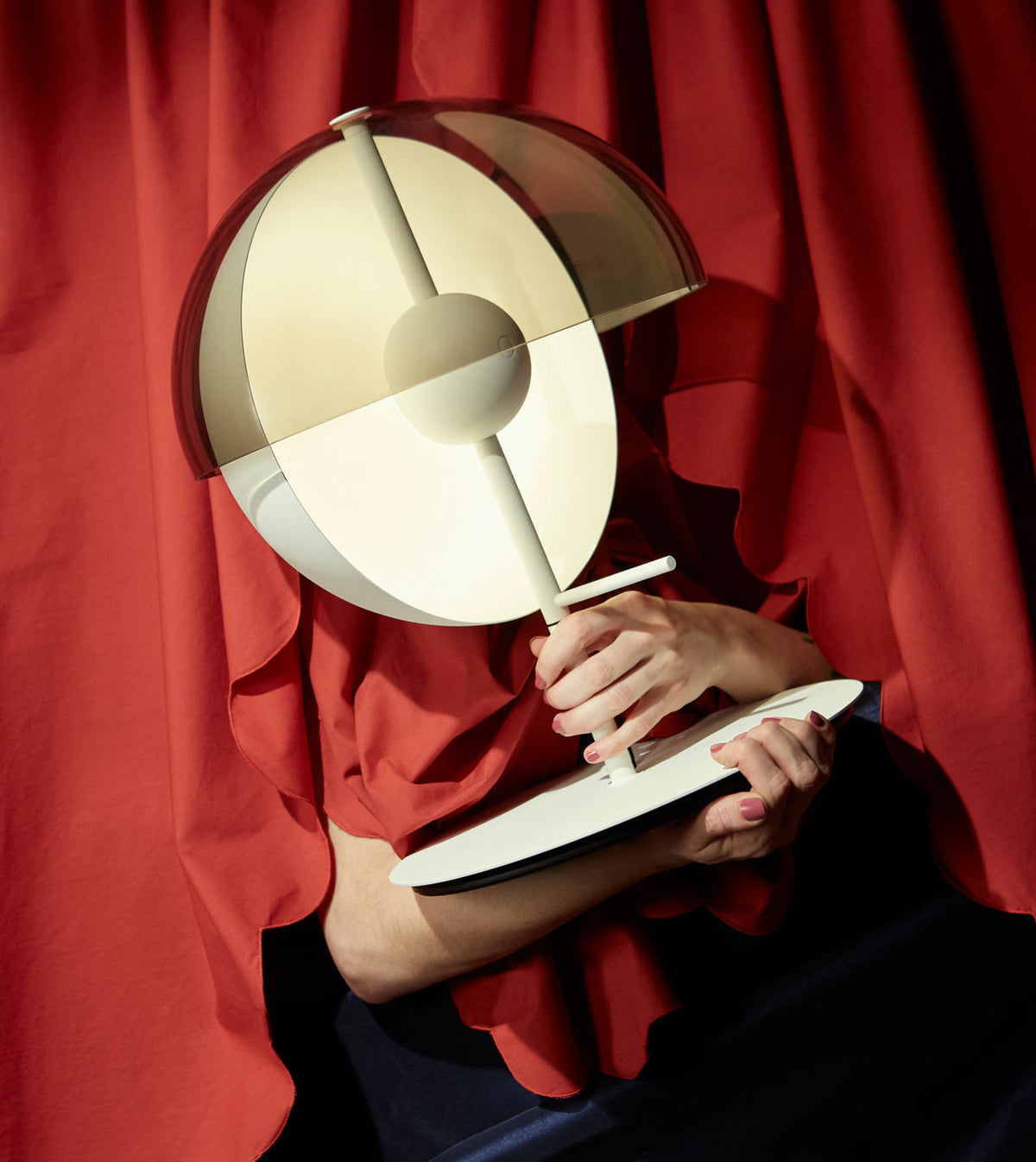 A white Clérachic table lamp is held in front of a red curtain, its dome and half-sphere tilted toward the camera.