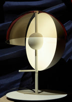 A white-based Clérachic table lamp with its shade opened out, lit against dark blue fabric.