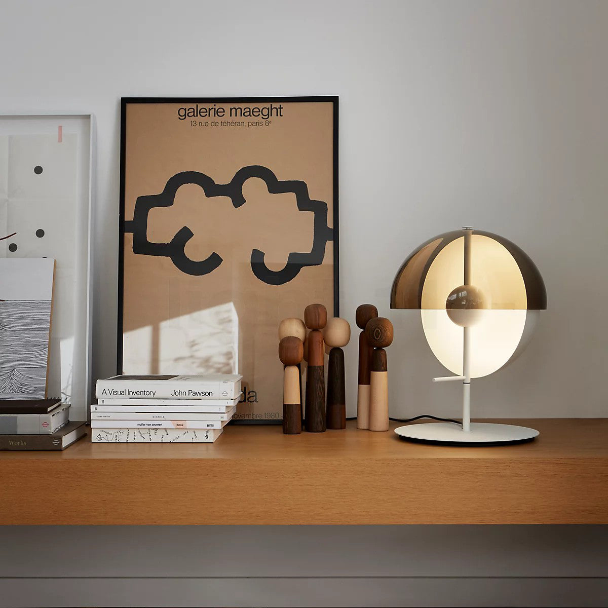 On a wooden shelf in front of a framed poster, books and carved wooden figures, a white-based Clérachic table lamp glows.