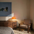 Clérachic's orange dome table lamp glows on a walnut sideboard, framed by a striped armchair in a sunlit bedroom.